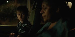 A young white boy is looking across to an older black woman in the front of a car